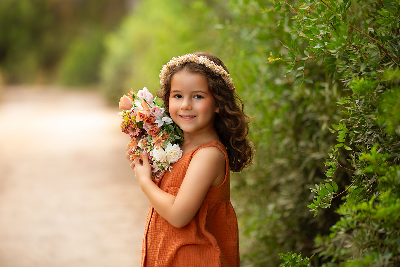 niña con vestida de color teja y corona de flores y sosteniendo un ramito de flores apoyado en su hombro, en su sesion infantil y familiar en la naturaleza en valencia